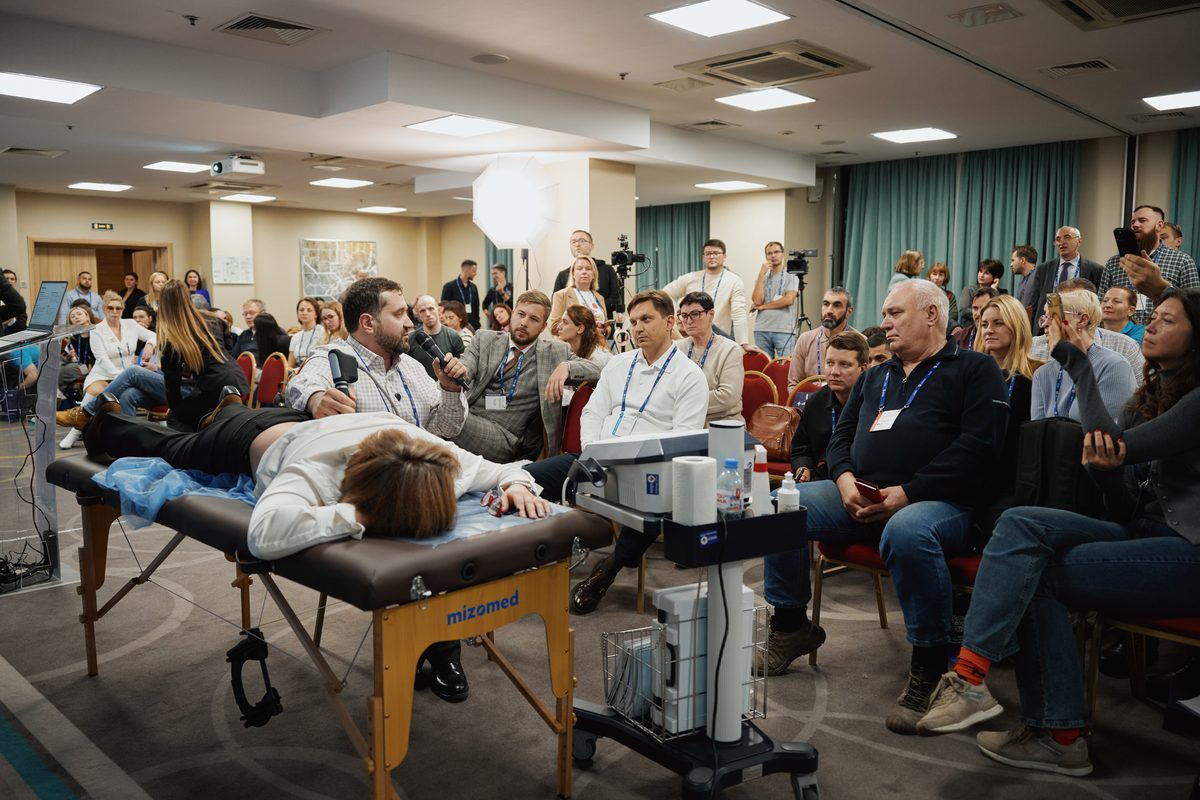 Демонстрация на конференции 4Clinic: спикер показывает методику у кушетки, участники смотрят вокруг